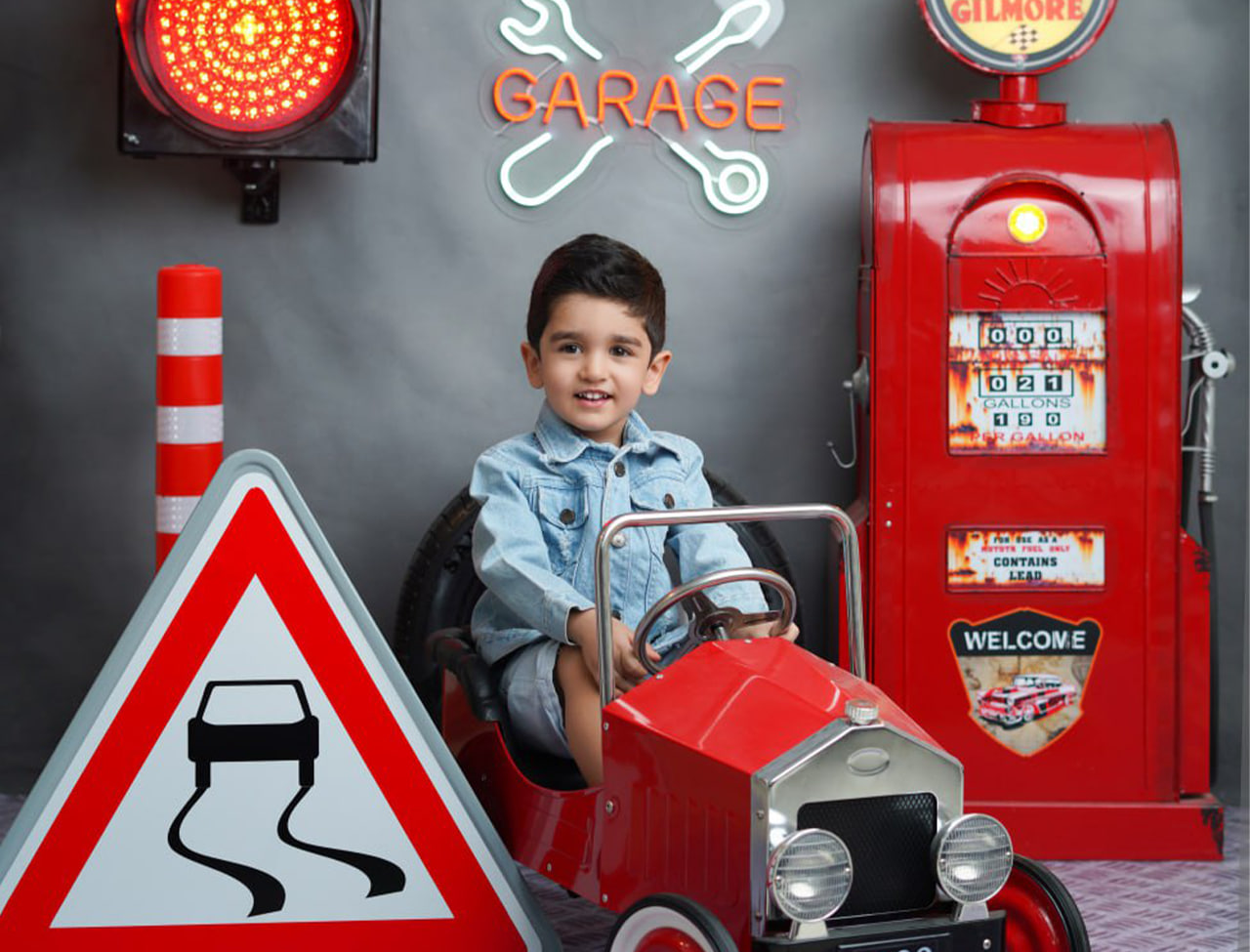 photo_2025-11-26_14-43-47 عکاسی کودک با دکور گاراژ قرمز در آتلیه تیرشات چیتگر، پسر بچه با لباس جین در ماشین کلاسیک قرمز